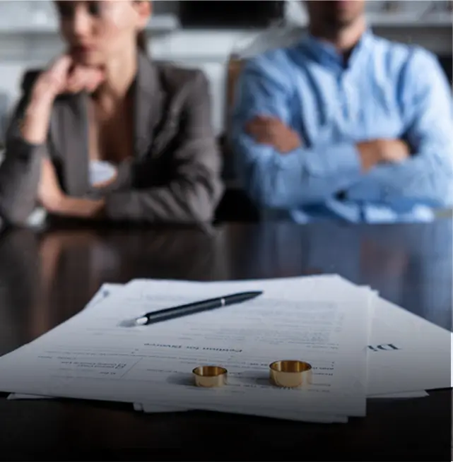 Two wedding rings on divorce papers with a pen, while a man and woman sit distantly in the background with crossed arms