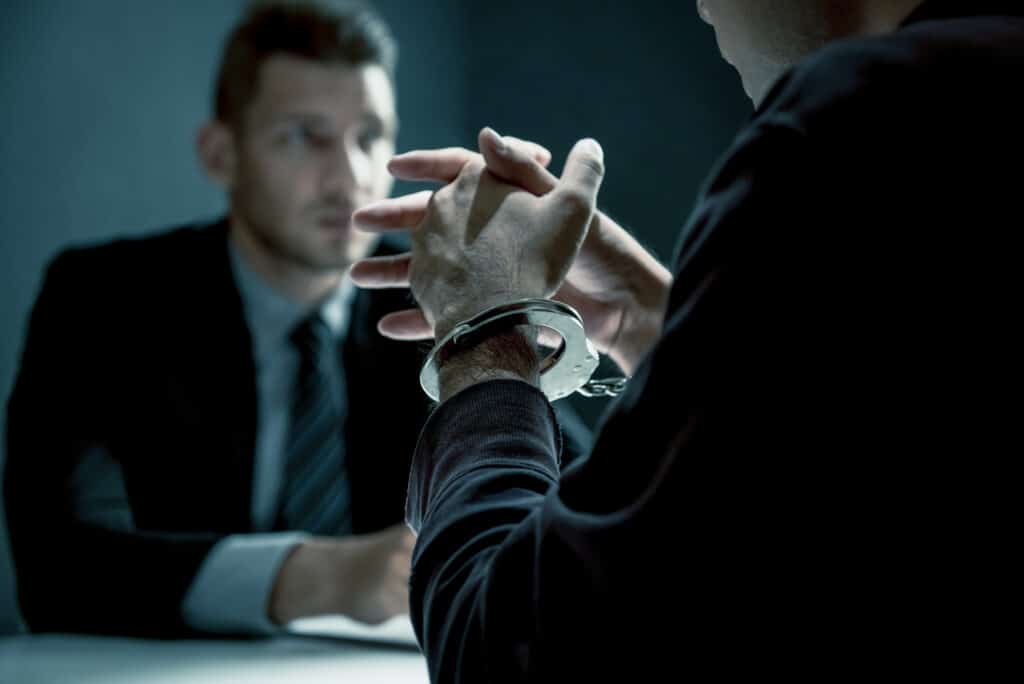 A man with handcuffs being interviewed in an interrogation room in front of a detective.